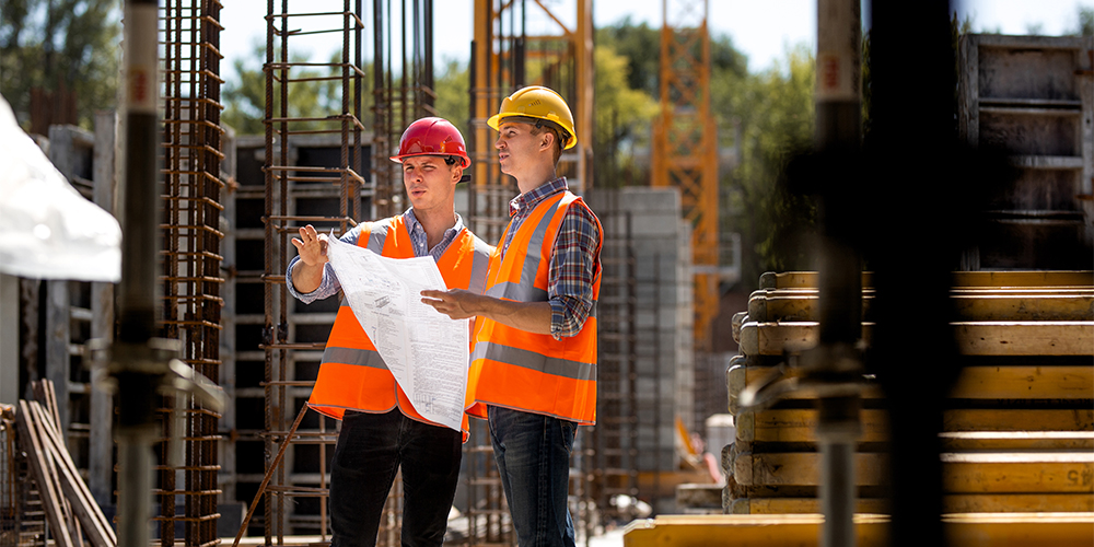 Two construction workers stand outside studying a paper Bill of Quantities.