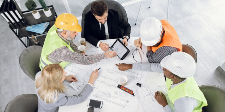 Photo of 5 different construction professionals of various types gathered around a meeting table which has construction drawings on it