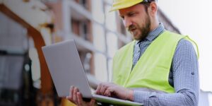 Man in a safety vest and hard hat stands on a jobsite looking at a laptop.