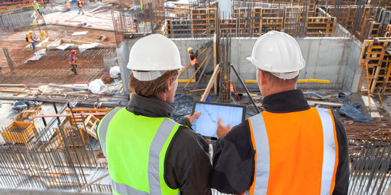 A construction site manager, wearing a hard hat (white for UK) and safety vest, stands holding a tablet next to a colleague. The screen displays construction management software with various metrics. The background shows an active construction site.