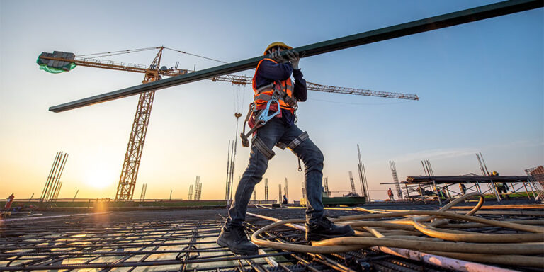 A construction worker in an high-vis vest carries a metal girder against a background of cranes and a construction site at sunset