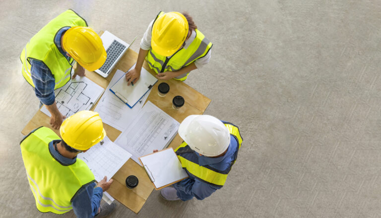 a view from above: 4 construction workers in hard hats and hi-vis study a construction management plan and other construction documents