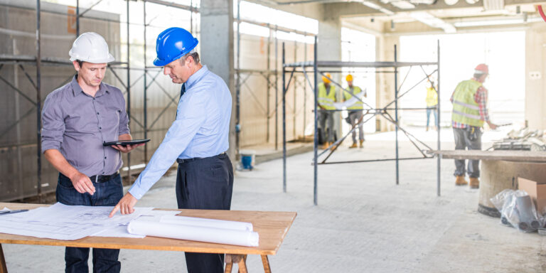 Architect and investor at a construction site talking and examining blueprints while using a digital tablet. Construction workers working in the background.