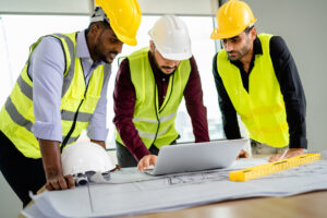 Team of Engineers diverse in safety suite discussing RIBA stages with blueprints and laptop on the desk at the workplace.