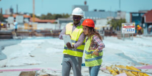 A young man engineer and his female colleague are looking at their tablet while standing on a construction site. A risk warning sign is in the background.