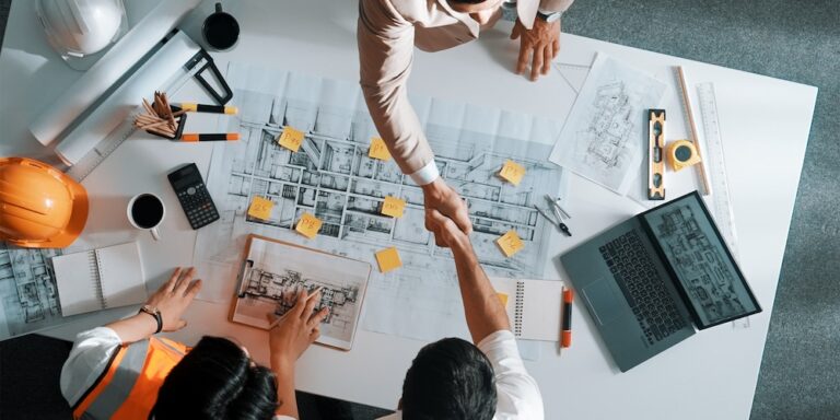 Two construction team members shaking hands over a table with construction drawings